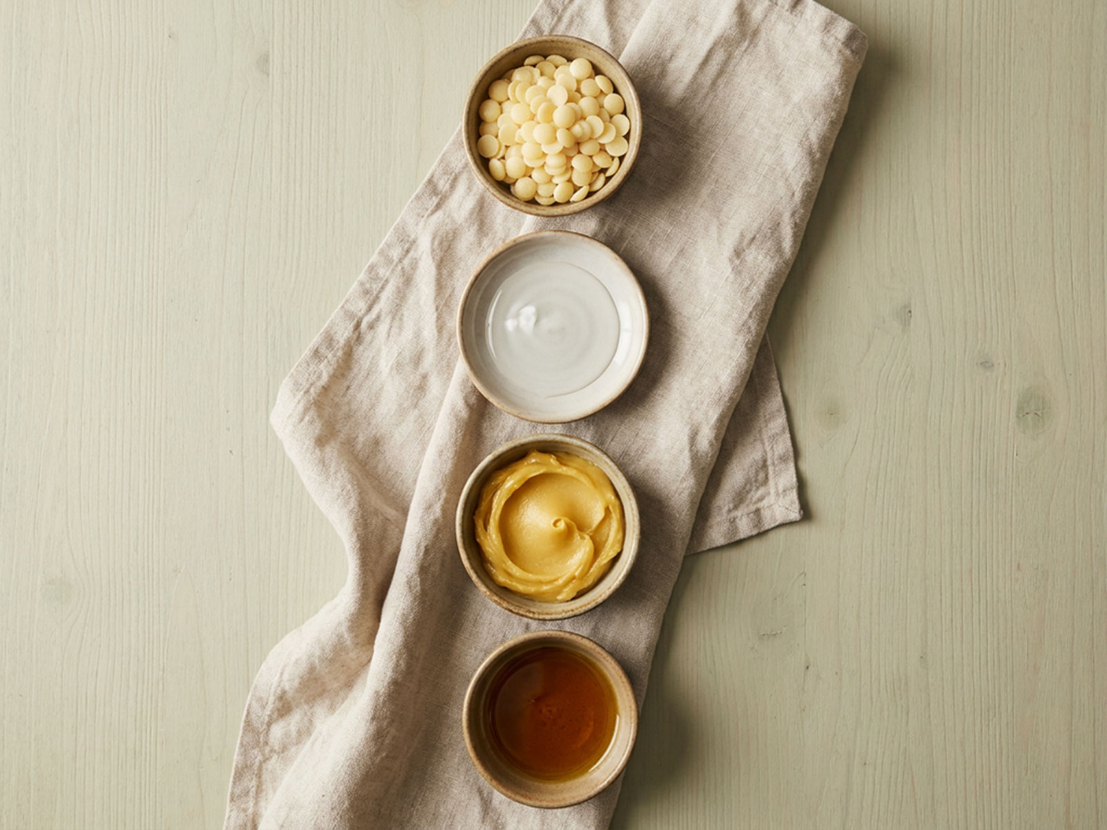 Overhead view of four small dishes on a linen cloth, holding pale wax pastilles, a clear emollient, a soft yellow butter and an amber oil