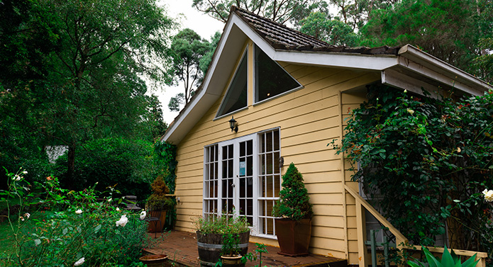 The Ferny Creek premises: a pale yellow weatherboard cottage with French doors and a gabled window, potted bay trees on the deck and a climbing vine up one wall, set among garden trees